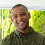 Meet You On The Mat A man in a green button-up shirt smiles while standing in front of a sunlit background with greenery visible through a window. Pilates & Dance