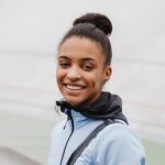 Meet You On The Mat A person is smiling at the camera, wearing a light blue jacket with a high collar and a black shoulder strap. Their hair is tied up in a bun. The background is blurred. Pilates & Dance