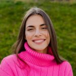 Meet You On The Mat A woman with long brown hair, wearing a bright pink turtleneck sweater, smiles against a grassy background. Pilates & Dance