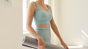 Meet You On The Mat Person in light blue activewear holding a rolled-up yoga mat in a bright, airy room, ready to meet you on the mat for Pilates. Pilates & Dance