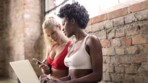 Meet You On The Mat Two women in sports bras stand next to a brick wall, one using a phone and the other working on a laptop, perhaps coordinating their next Pilates session or planning to Meet You On The Mat. Pilates & Dance