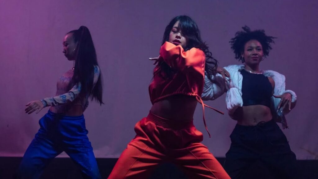 Meet You On The Mat Three dancers, dressed in varied outfits, perform synchronized dance moves on a dimly lit stage with purple lighting, seamlessly blending elements of Pilates. Pilates & Dance
