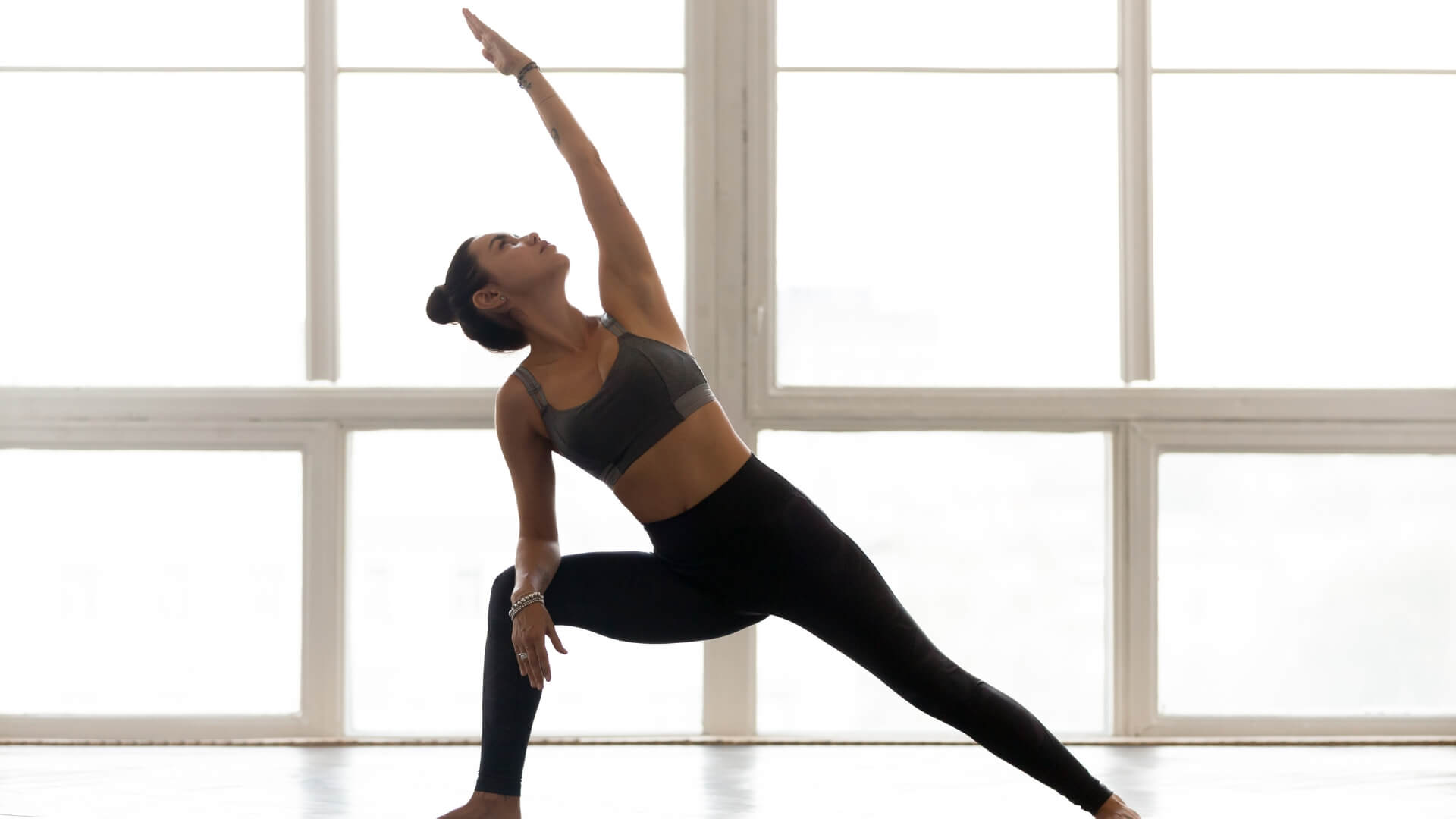 Meet You On The Mat A person in athletic wear is performing a yoga pose with one arm raised and the other hand resting on the floor inside a bright room with large windows, seamlessly blending the grace of dance into their practice. Pilates & Dance