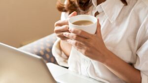 Meet You On The Mat A person wearing a white shirt holds a cup of coffee in both hands while sitting next to an open laptop, ready to start their day with some Pilates. Pilates & Dance