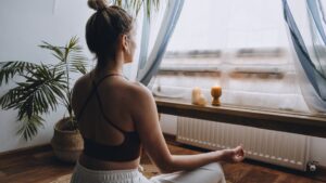 Meet You On The Mat A woman sits cross-legged on a wooden floor in front of a window, meditating with candles placed on a low table beside her. She is wearing a sports bra and light-colored pants, creating an ambiance that whispers serenity as if to say, "Meet You On The Mat. Pilates & Dance