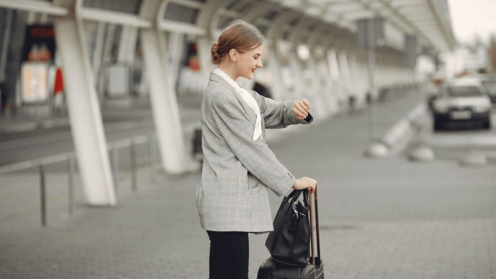 Meet You On The Mat A woman in a gray blazer stands outdoors checking her watch, holding the handle of a black suitcase in her other hand, ready for the next adventure. Pilates & Dance