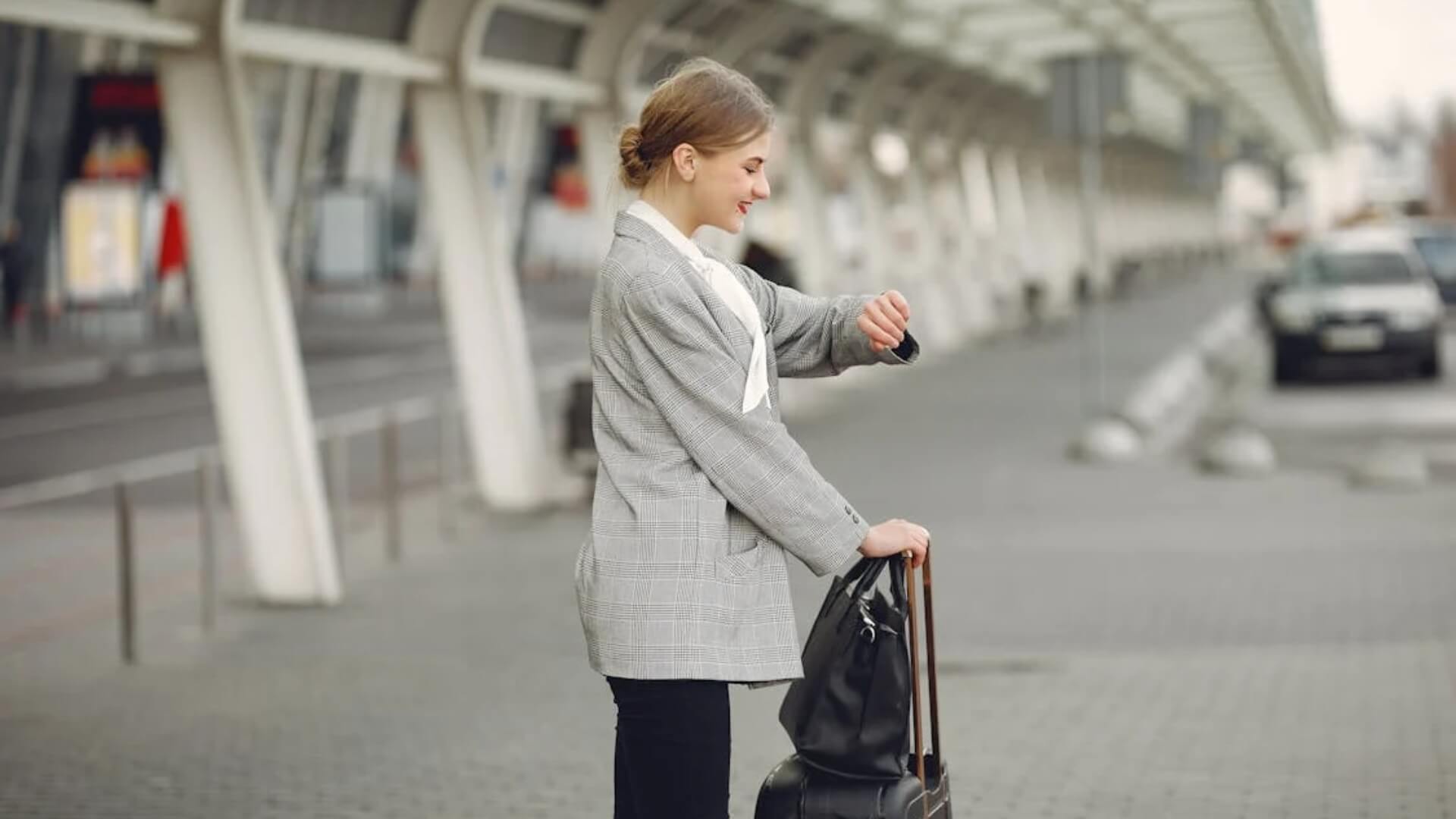 Meet You On The Mat A person in a gray blazer and black pants checks their watch while holding a black bag at a modern, covered outdoor location, possibly anticipating their upcoming Pilates session. Pilates & Dance