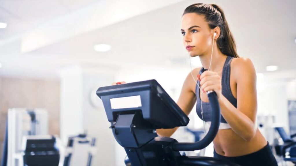 Meet You On The Mat A woman in athletic wear uses an elliptical machine in a gym while listening to music through earphones, imagining the beat as a dance routine. Pilates & Dance