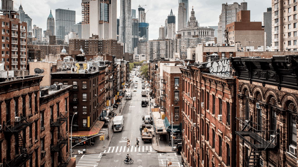 Meet You On The Mat A city street with cars, bicycles, and pedestrians is lined with brick buildings, with tall skyscrapers visible in the background. Pilates & Dance