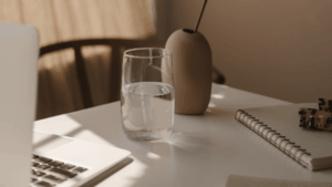 Meet You On The Mat A glass of water sits on a white desk next to a laptop, a notebook, a pen, and a beige vase in soft natural light. Pilates & Dance