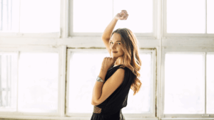 Meet You On The Mat A woman with long hair in a black dress poses indoors in front of large windows, with one arm raised and the other bent near her chin. Pilates & Dance