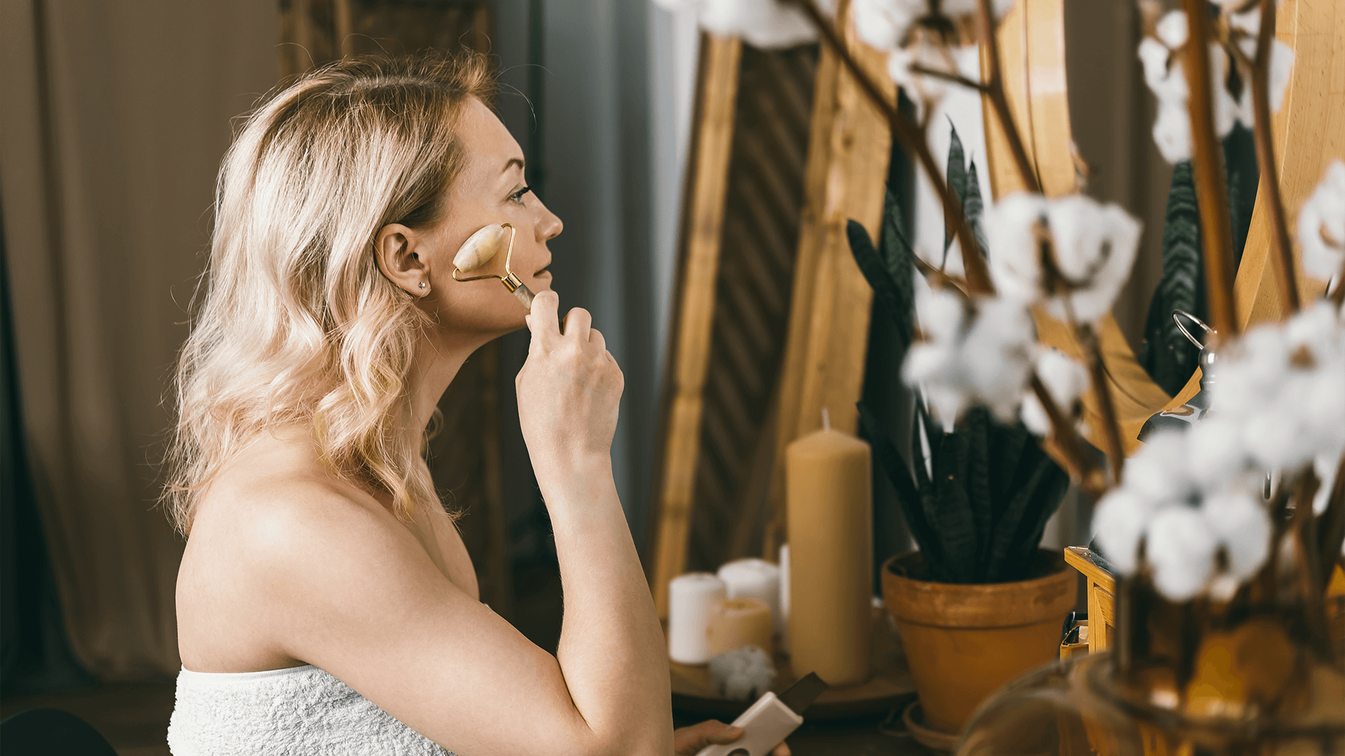 Meet You On The Mat A woman in a towel uses a facial roller on her cheek while sitting at a vanity table with candles, plants, and decorative items. Pilates & Dance