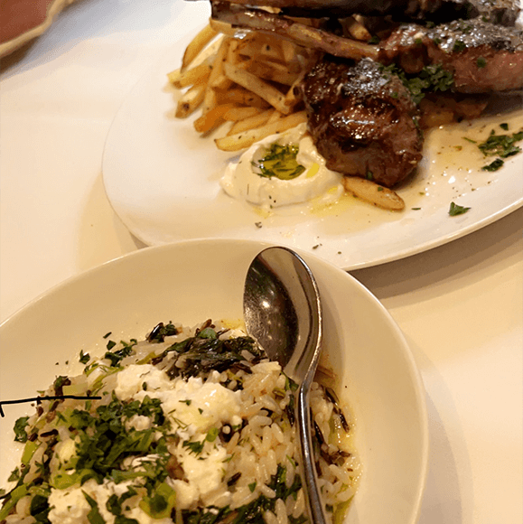 Meet You On The Mat A bowl of orzo pasta topped with herbs and cheese sits in front of a plate of grilled lamb chops, fries, and a dollop of sauce on a white tablecloth. Pilates & Dance