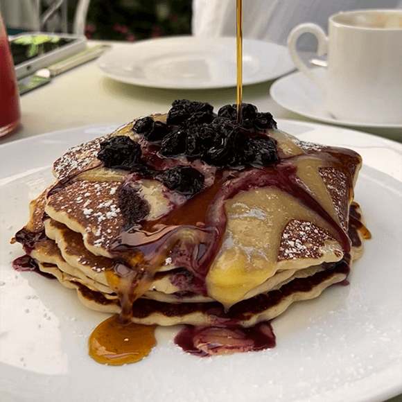 Meet You On The Mat A stack of pancakes topped with blueberries and powdered sugar is being drizzled with syrup, served on a white plate with a cup and saucer in the background. Pilates & Dance