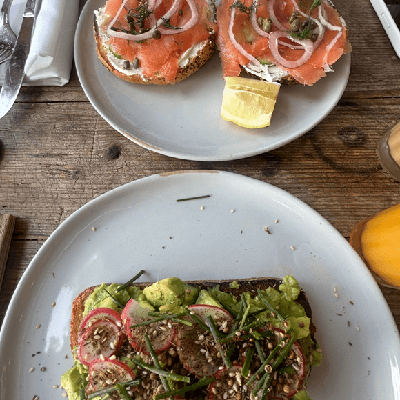 Meet You On The Mat Two plates on a wooden table: one with smoked salmon toast and lemon, the other with avocado toast topped with radish, seeds, and greens. A glass of orange juice is nearby. Pilates & Dance
