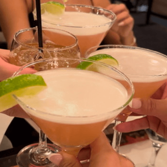 Meet You On The Mat Four people clink cocktails together, with three pink drinks garnished with lime and one brown drink with ice and a straw. Pilates & Dance