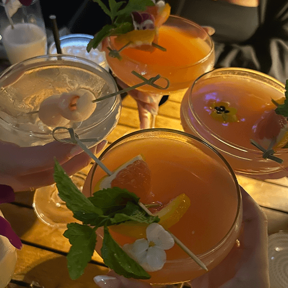 Meet You On The Mat Four hands hold colorful cocktails garnished with fruit, flowers, and herbs over a wooden table in a dimly lit setting. Pilates & Dance