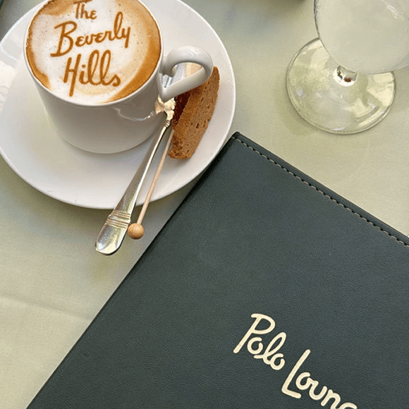 Meet You On The Mat A cappuccino with "The Beverly Hills" written in foam sits on a saucer with a biscotti, next to a Polo Lounge menu and a glass of water on a table. Pilates & Dance. LA Food Favs