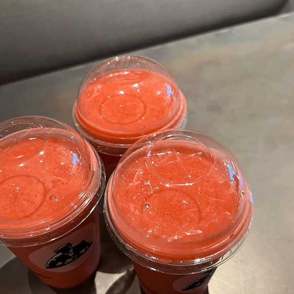 Meet You On The Mat Three clear plastic cups filled with bright red slushy drinks, each covered with a domed lid, placed on a metallic surface. Pilates & Dance. New York City Food Favs