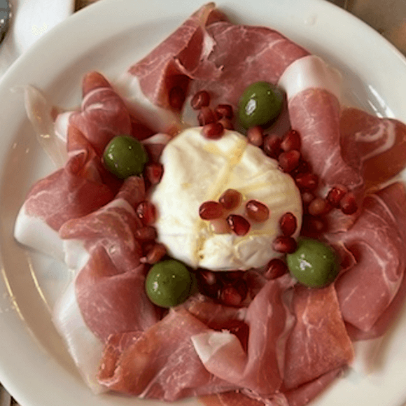 Meet You On The Mat Plate with slices of prosciutto, a ball of burrata cheese in the center, green olives, and pomegranate seeds. Pilates & Dance