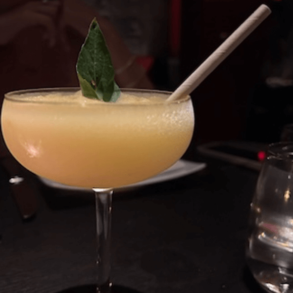 Meet You On The Mat A yellow cocktail in a coupe glass garnished with a green leaf and served with a paper straw, placed on a dark table. Pilates & Dance. New York City Food Favs