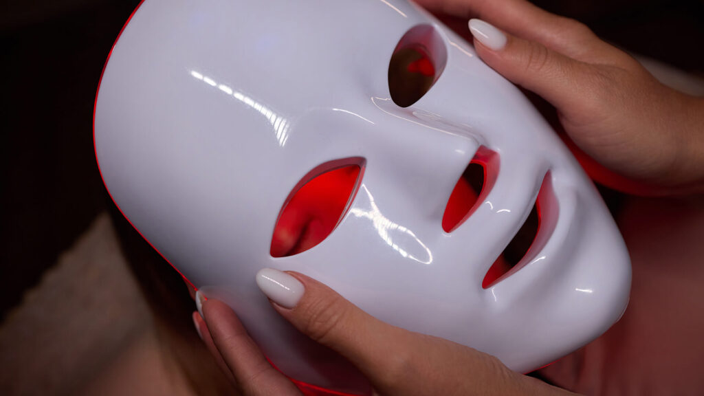 Meet You On The Mat A person holding a white LED light therapy mask with red light glowing through the eye and mouth openings. Pilates & Dance