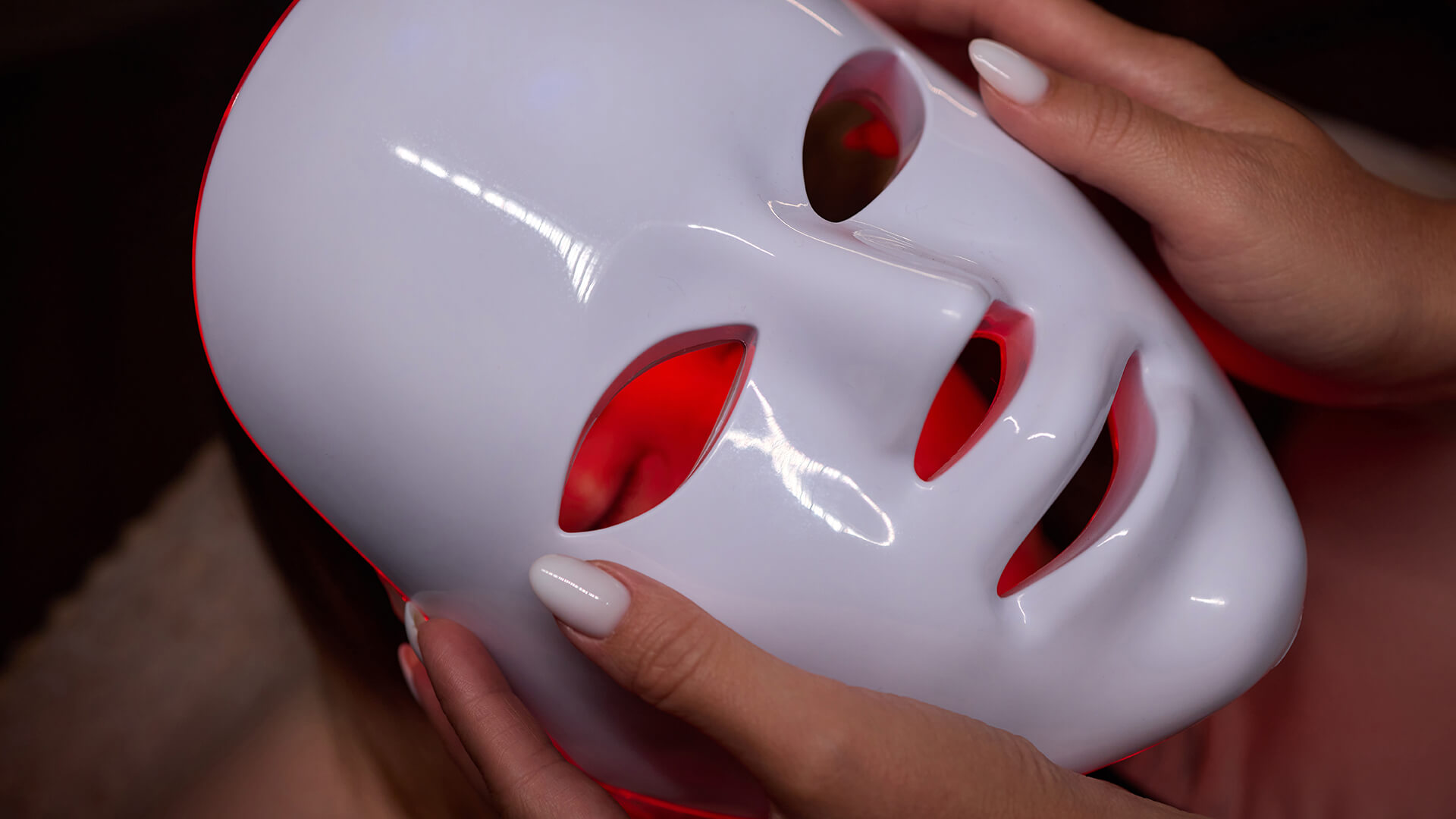 Meet You On The Mat A person holding a white LED light therapy mask with red light glowing through the eye and mouth openings. Pilates & Dance