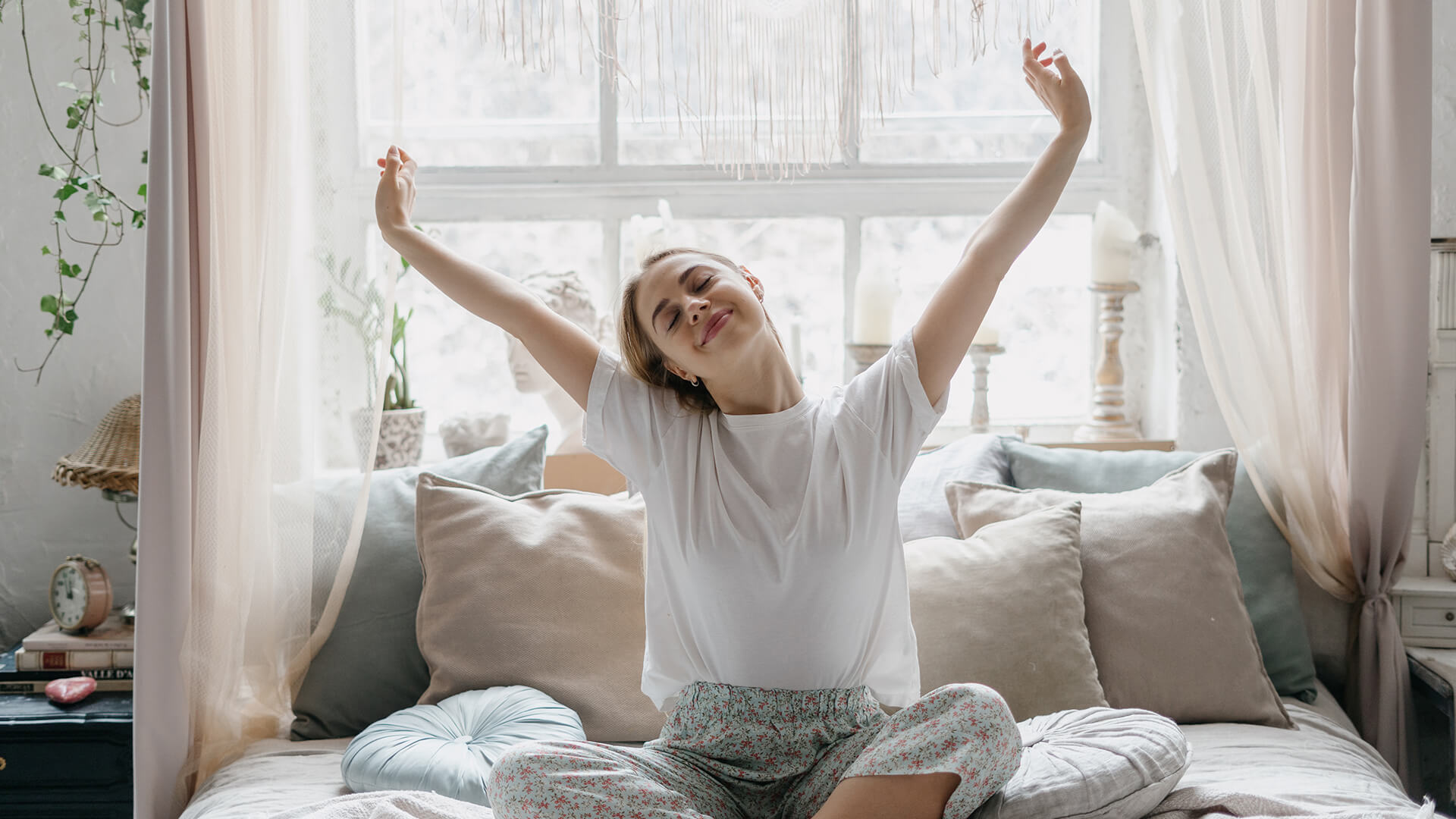 Meet You On The Mat A person sits cross-legged on a bed, stretching their arms upward and smiling, in a bright room with large windows and pillows. Pilates & Dance